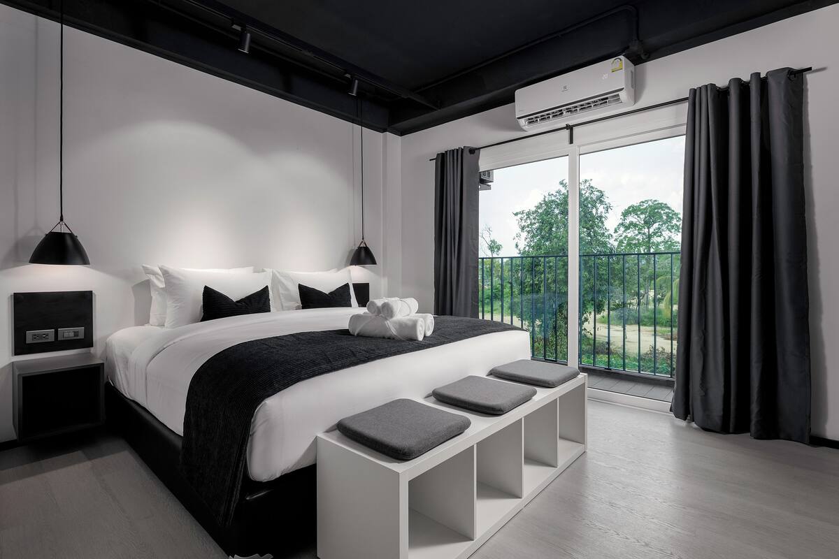 A modern bedroom with a large bed, bedside tables, a bench, and a view of greenery through a balcony.