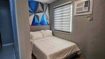 Un dormitorio con una cama, un mural geométrico en la pared, una ventana con persianas y un equipo de aire acondicionado instalado en la pared.