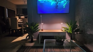 A living room with a flat-screen TV mounted on the wall, a glass coffee table, and two potted plants.