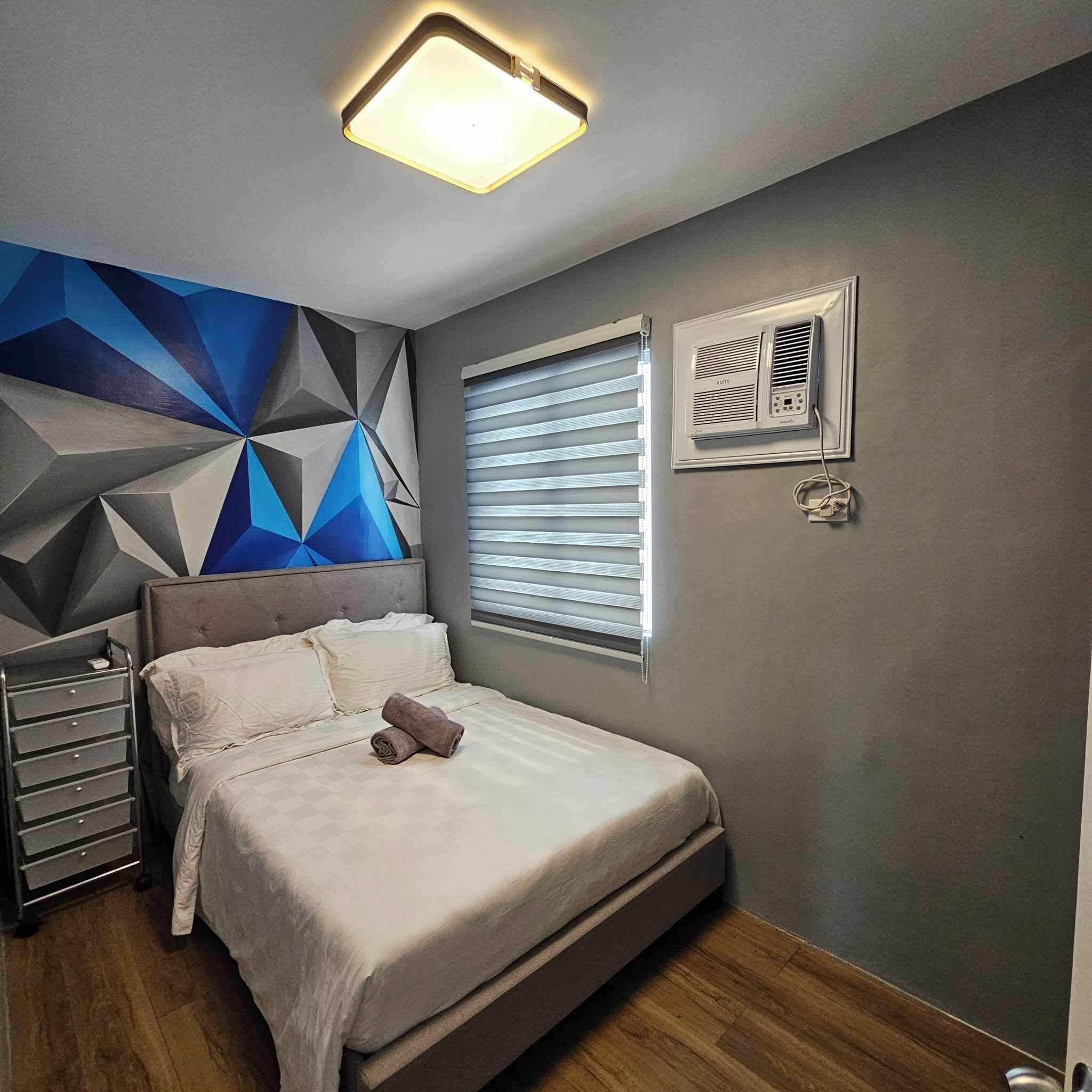 A modern bedroom with a geometric wall design, a bed with white bedding, a bedside table, and a wall-mounted air conditioning unit.