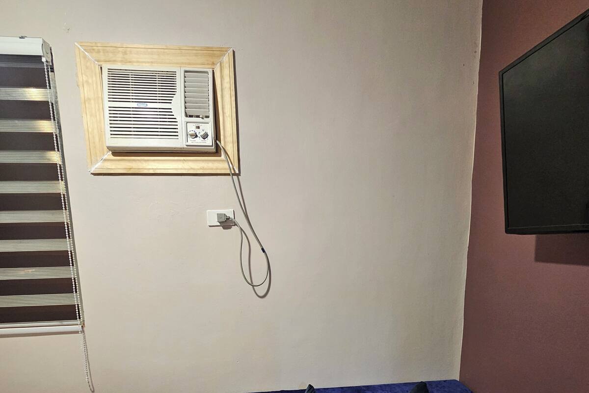 A bedroom with a single bed, a window with blinds, an air conditioning unit, and a wall-mounted TV.
