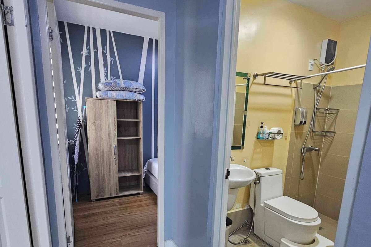 A compact bathroom with a toilet, sink, and shower. There is a wooden wardrobe and a bed in the adjacent room.