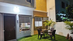 A two-story building with a patio area featuring a table and chairs, artificial grass, and a stone pathway.