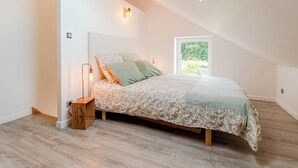 A bedroom with a sloped ceiling, a bed with patterned bedding, a bedside lamp, and a window with a view of greenery.