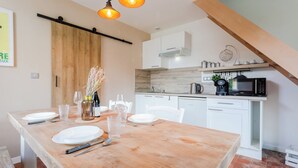 A dining area with a wooden table set for four, white chairs, and a kitchen in the background with white cabinets and a microwave.