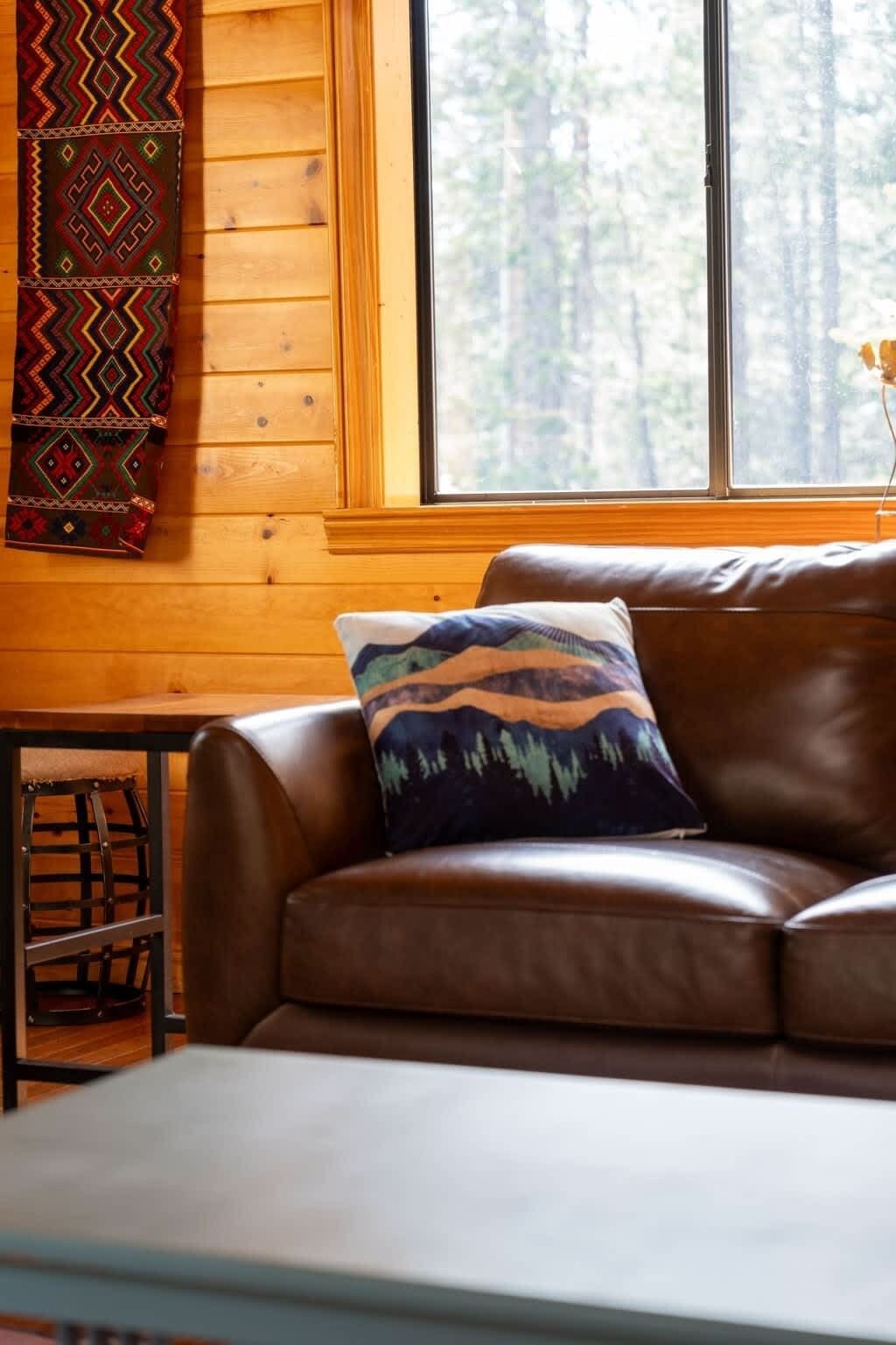 A brown leather sofa with a patterned pillow, a wooden table, and a window with a view of trees.