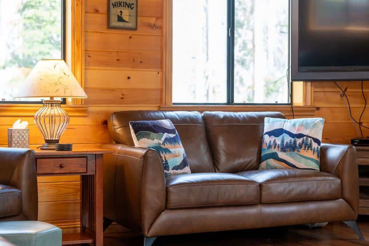 A leather sofa with two decorative pillows, a wooden side table with a lamp, and a television mounted on the wall.