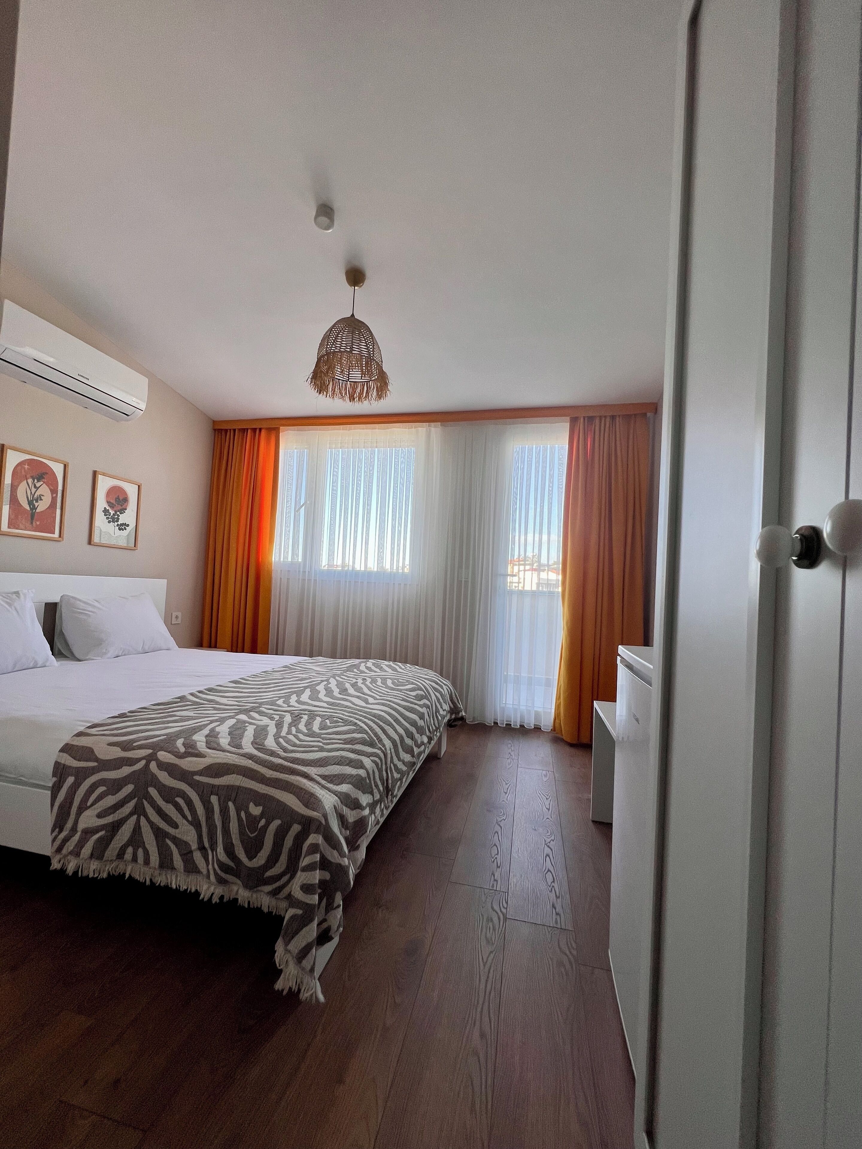A hotel room with a bed, a bedside table, an air conditioning unit, a hanging light fixture, and a window with curtains.