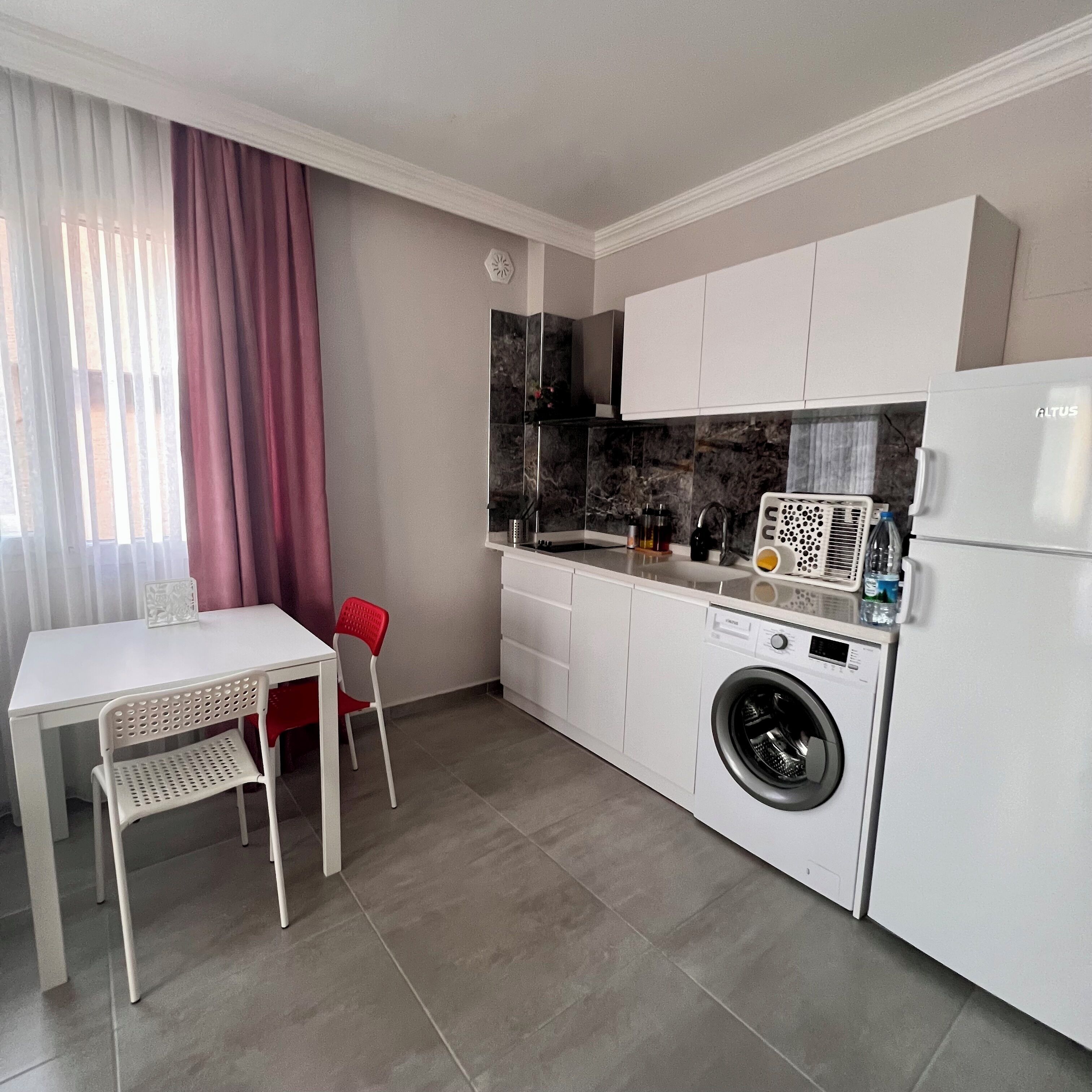 Una cocina compacta con lavadora, una mesa con sillas y un ventanal con cortinas.