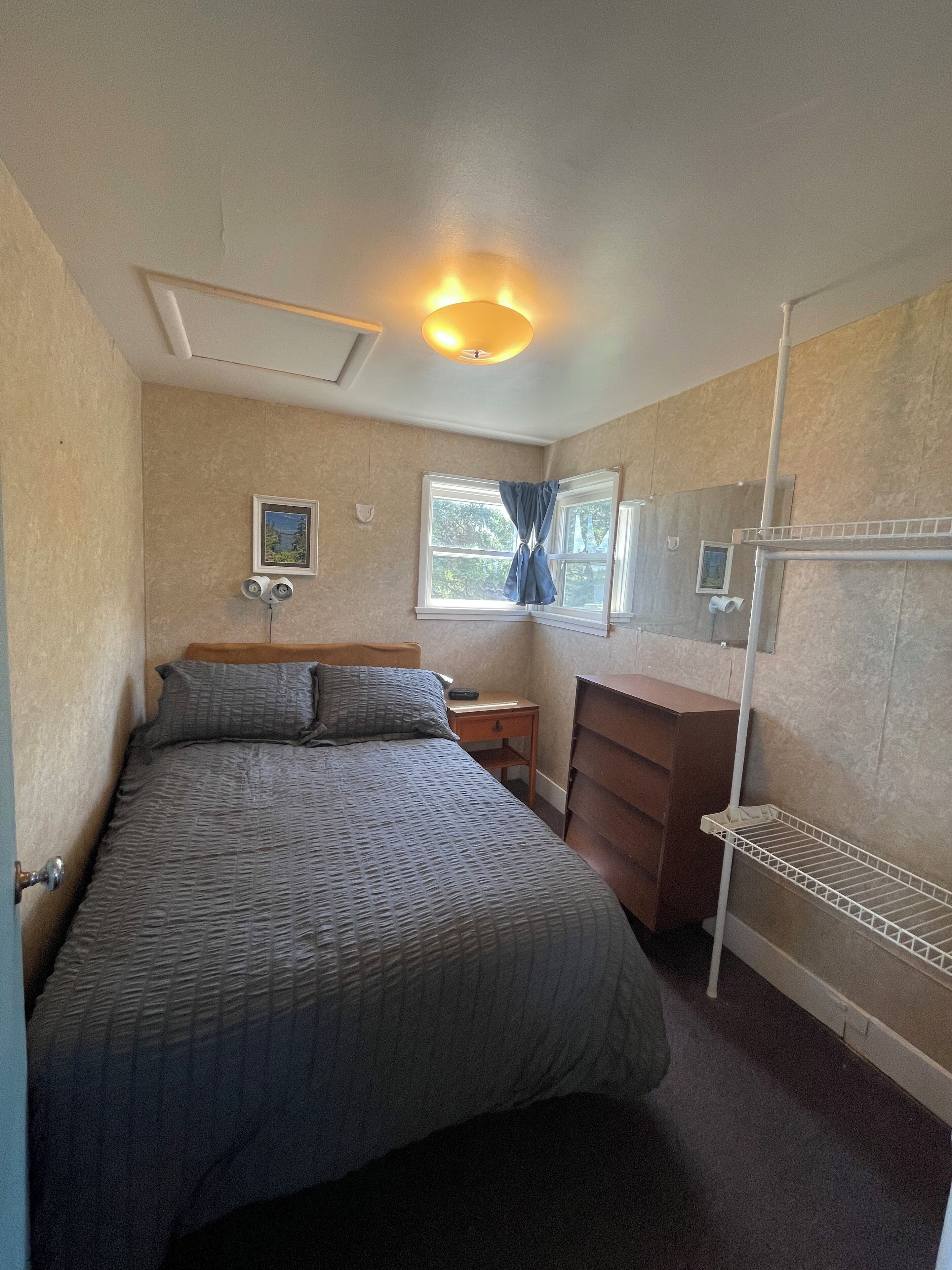 Un dormitorio con cama, cómoda, mesita de noche, estante y ventana con cortinas.