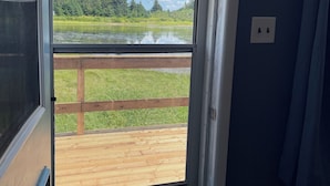A view through a door with a wooden deck, a grassy area, and a lake with trees in the background.