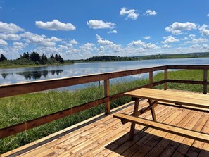 A wooden deck with a bench overlooking a calm lake, surrounded by grassy areas and trees. - Secluded Waterfront Cottage with Kayaks  (Afton Station)
