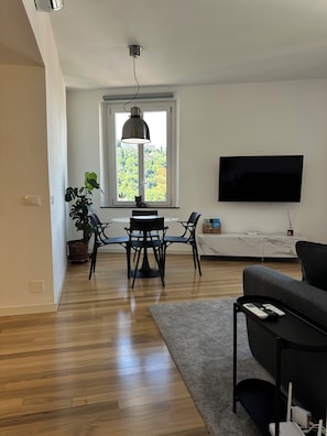 A modern living room with a flat-screen TV, a dining area with a table and chairs, a grey sofa, a potted plant, and a large window with a view of trees. - Casa di May Acquasanta - Apartment B (La Spezia)
