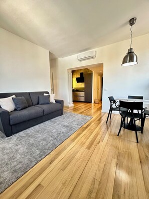 A modern living room with a grey sofa, a dining table with chairs, wooden flooring, and a kitchen visible in the background. - Casa di May Acquasanta - Apartment B (La Spezia)