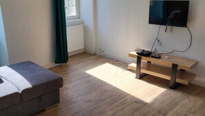 A room with a wooden floor, a grey sofa, a wooden bench, a TV mounted on the wall, and a window with curtains. - Large apartment 1st floor (Saint-Pol-de-Léon)