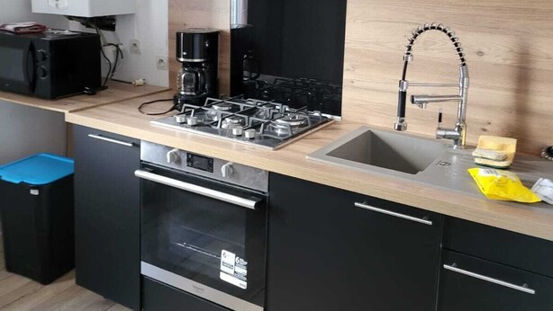 A compact kitchen with a gas stove, oven, and sink. Red cabinets and a white range hood are visible. - Large apartment 1st floor (Saint-Pol-de-Léon)