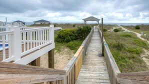 Une promenade en bois menant à des cabanes de plage et à un kiosque, le tout entouré de dunes et de végétation.