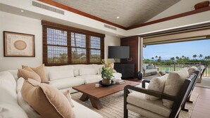 A spacious living room with a large sofa, a coffee table, and a television. There is a large window with bamboo blinds, and a view of palm trees and the ocean.
