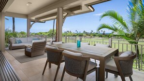 A covered outdoor area with a wooden table and wicker chairs, a sofa, and a view of the ocean and palm trees.