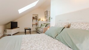 A bedroom with a sloped ceiling, a bed with patterned bedding, a television on a dresser, and shelves with books and decorative items.
