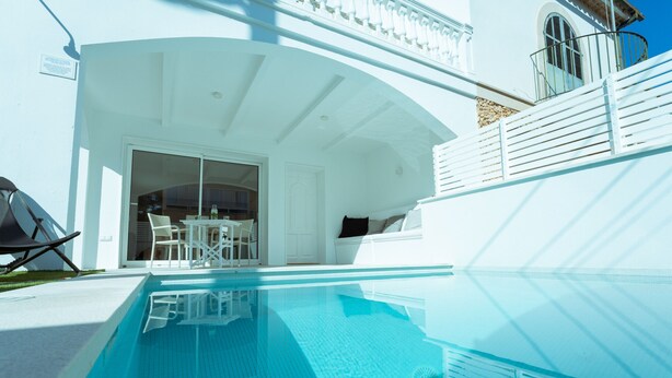 A clear blue swimming pool with a sunlit patio area, a white outdoor seating arrangement, and a white building with a balcony.