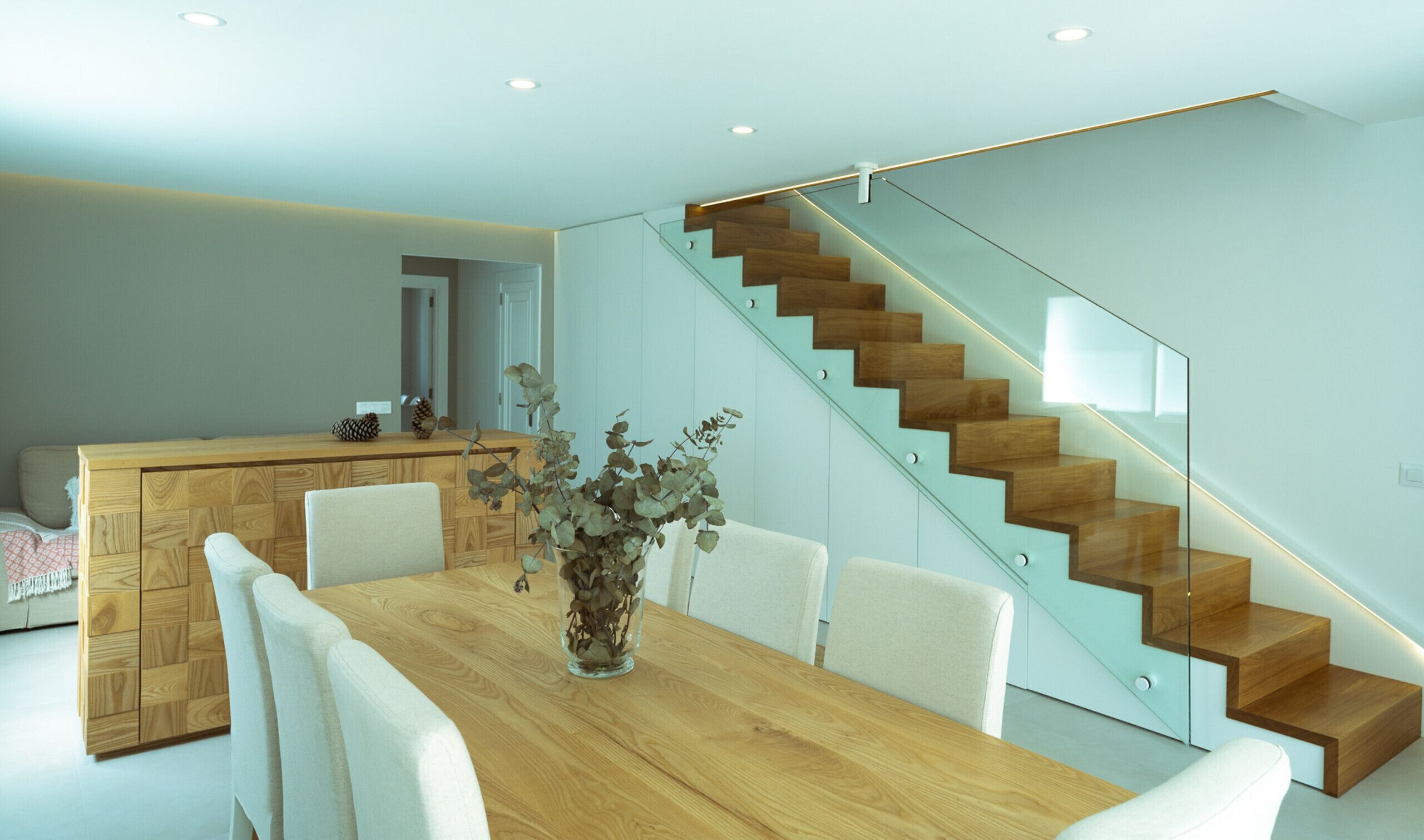 A modern kitchen with a wooden cabinet, a dining table with chairs, and a glass staircase.