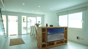 A modern living room with a wooden entertainment unit, a flat-screen TV, and a dining area with white chairs.