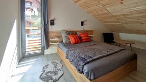 Un dormitorio con techo inclinado, una cama con almohadas grises y rojas, una mesita pequeña y una ventana con cortinas.