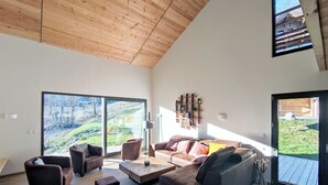 Un salón amplio con techo de madera alta, ventanas grandes que ofrecen vistas a una ladera, y una zona de estar acogedora con sofás y otomanas.