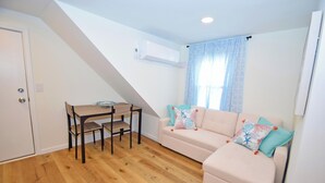 Een kleine, goed verlichte kamer met een houten vloer, een beige bank met kleurrijke kussens, een klein tafeltje met een stoel, en een raam met lichtblauwe gordijnen.