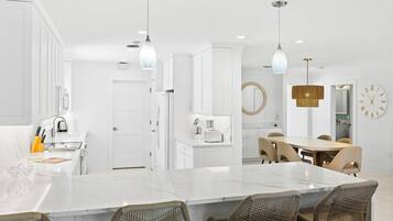Une cuisine moderne avec des meubles blancs, un îlot en marbre, des chaises en osier et des luminaires suspendus.