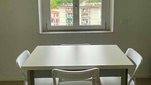 Une pièce avec une table blanche et deux chaises blanches, une fenêtre donnant sur des bâtiments et de la verdure.