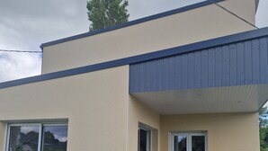 A modern building with a flat roof, a covered patio area with a table and chairs, and a small garden area with a bench.