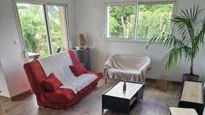 A spacious living room with a red sofa, a white armchair, and a coffee table. There are two windows providing natural light and a view of greenery outside.