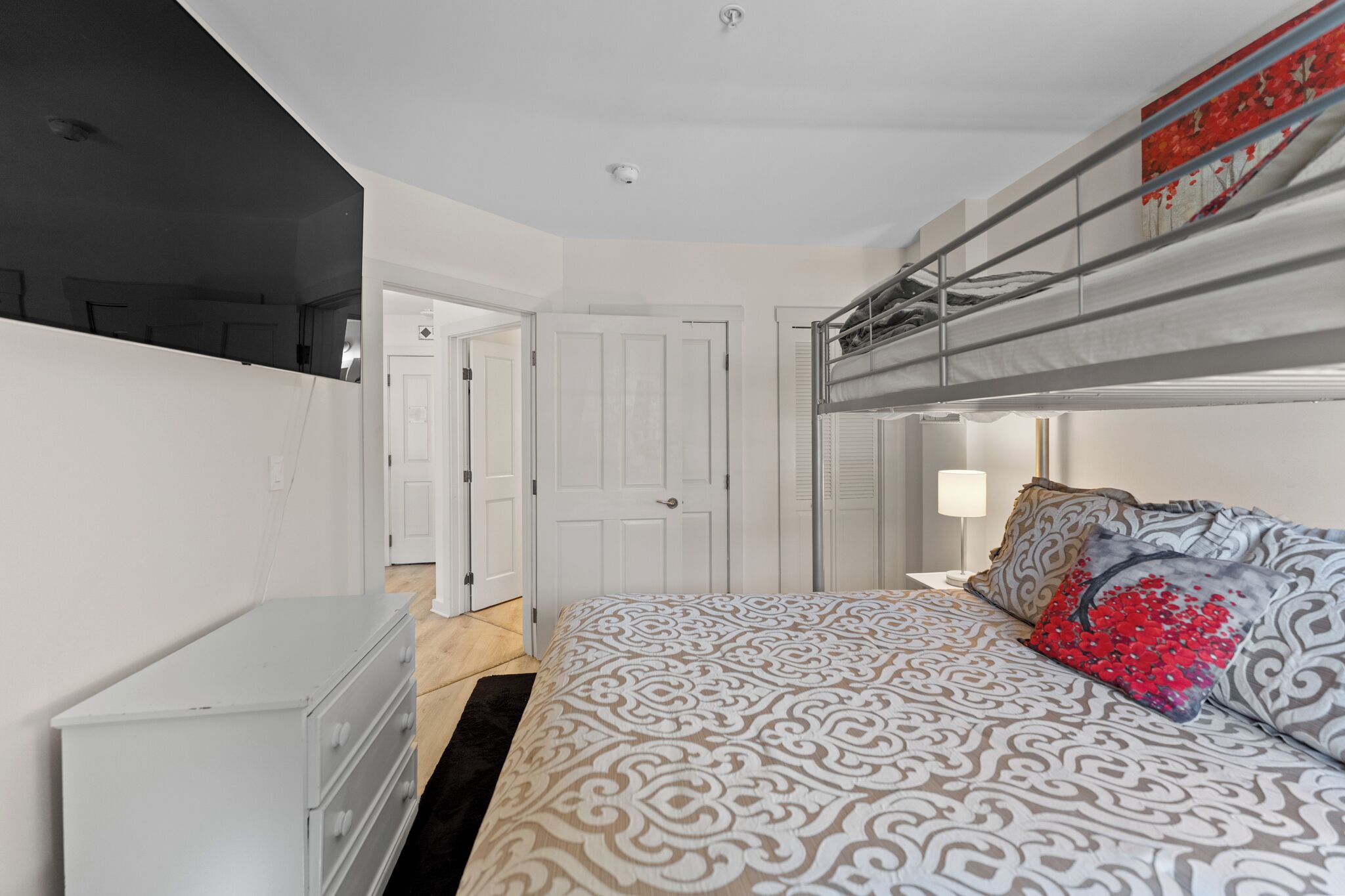A bedroom with a bunk bed, a dresser, a bedside lamp, and a door leading to another room.