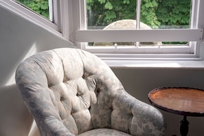 A tufted armchair with a floral pattern, positioned next to a window with a white frame, and a small round wooden table beside it.