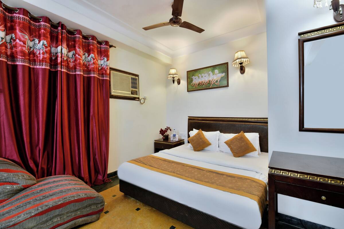 Une chambre d’hôtel avec un lit, une table de chevet, une lampe, un ventilateur de plafond et une fenêtre avec des rideaux.