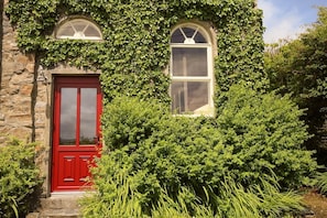 A stone building with a red door and two arched windows, surrounded by lush greenery. - Old School House, Clifden (Clifden)