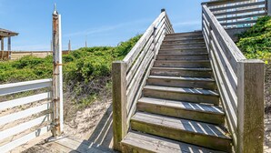 A wooden staircase leading to a beach area with a white fence and a wooden structure in the background. - The Minor Manor - NH MP 11.5, Historic Oceanfront, Dunetop Deck, YR Flex Stays (Nags Head)