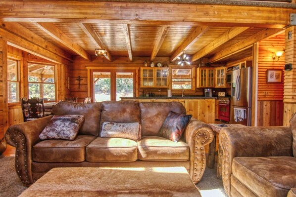 A cozy cabin interior with a leather sofa, a wooden dining table, and a kitchen area with a refrigerator and cabinets.