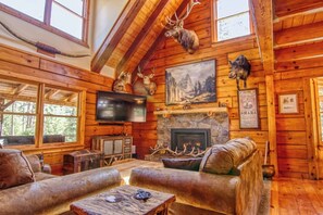 A log cabin interior with a stone fireplace, leather couches, mounted animal heads, and a television.