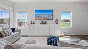 Un salon spacieux doté d’un grand téléviseur à écran plat fixé au mur, d’un canapé confortable et offrant une vue sur la plage grâce à de grandes fenêtres.