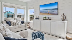 Un salon doté d’un grand téléviseur à écran plat fixé au mur, d’un canapé blanc agrémenté de coussins à motifs, d’une petite table d’appoint et offrant une vue sur la plage grâce à de grandes fenêtres.