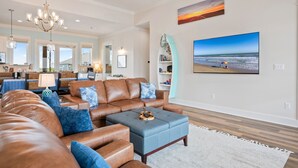 Een ruime woonkamer met een grote bruine leren bank, een blauwe poef en een kroonluchter. Op de muur hangt een groot schilderij van een strandlandschap.