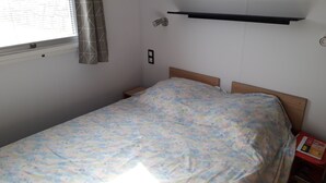A small, single-bed room with a wooden headboard, a bedside table, a wall-mounted shelf, and a window with curtains. - 2 bedroom mobile home in saint-raphaã "l (Saint-Raphaël)