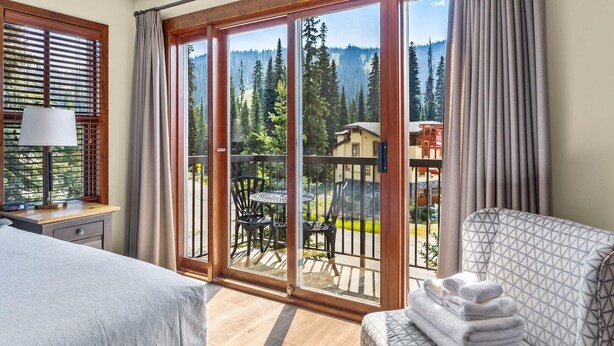 A hotel room with a bed, a chair, a table, and a balcony with a view of trees and buildings.