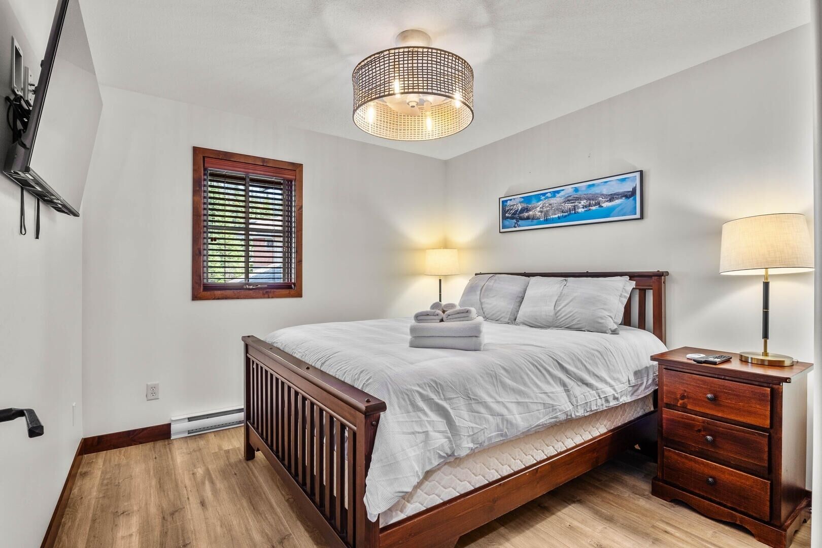 A bedroom with a wooden bed, a nightstand, a lamp, a window with blinds, and a painting on the wall.
