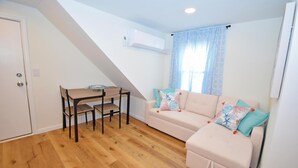 A small, well-lit room with a wooden floor, a sofa with colorful cushions, a small table with chairs, and a window with light blue curtains.