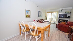 A dining area with a wooden table and white chairs, a red sofa, and a television on a shelf. - Park Place 101 2 Bedroom Condo by RedAwning (Ocean City)
