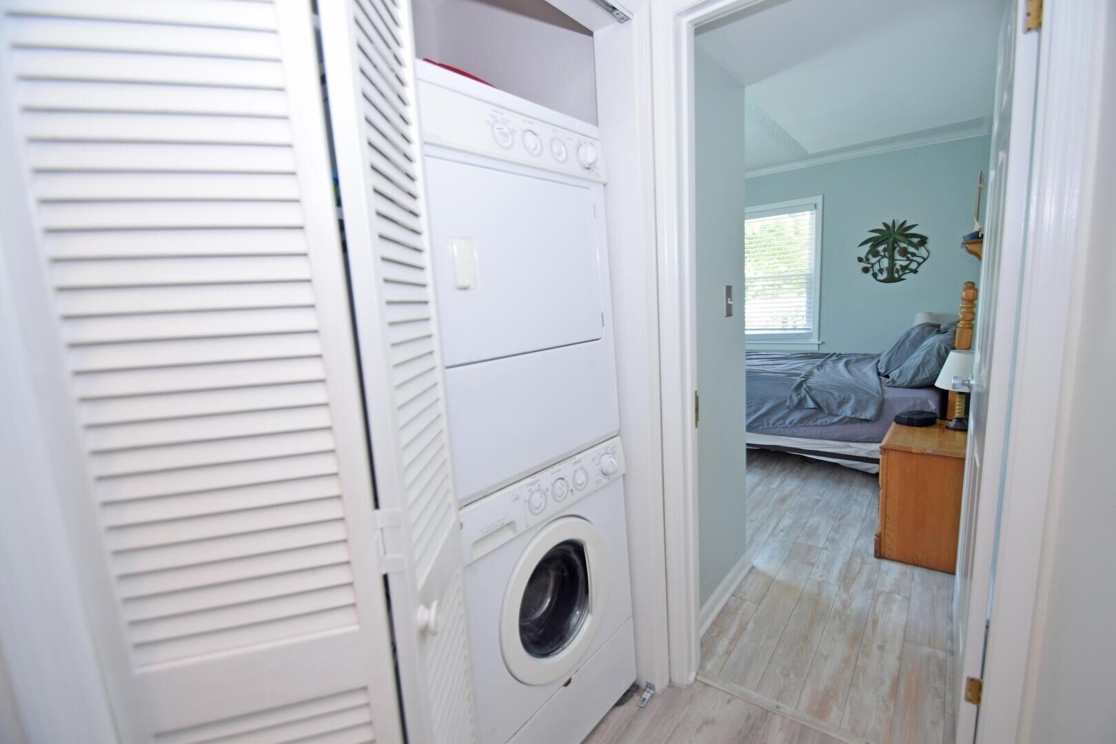 Una lavandería con lavadora y secadora de carga frontal, piso de madera y una puerta que da a un dormitorio con cama y mesita de noche.